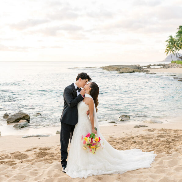 A bride and groom kiss on a sandy beach at sunset, with the ocean and palm trees in the background. The bride holds a colorful bouquet.