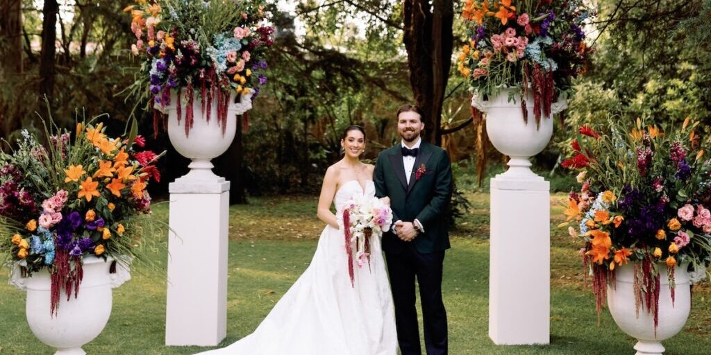 A bride and groom stand outdoors between two large floral arrangements on pillars, posing for a wedding photo.
