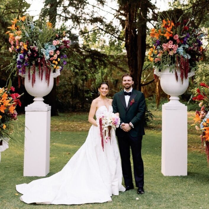 A bride and groom stand outdoors between two large floral arrangements on pillars, posing for a wedding photo.