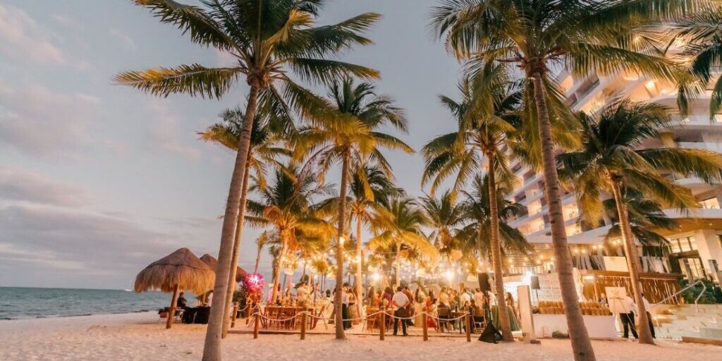 Outdoor beach event at sunset with people gathered under palm trees and string lights, near a thatched hut and a multi-story building.
