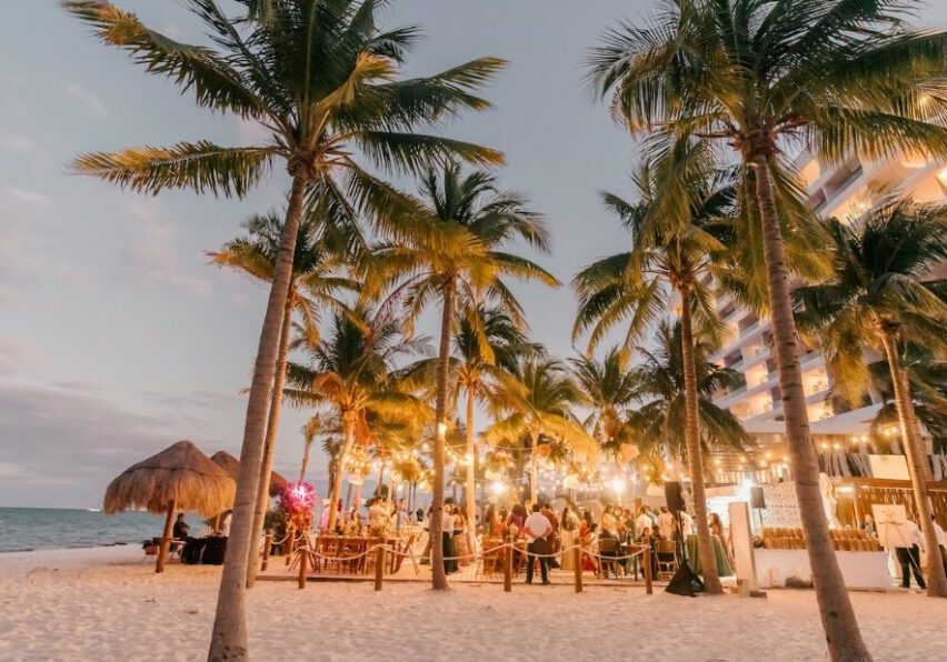 Outdoor beach event at sunset with people gathered under palm trees and string lights, near a thatched hut and a multi-story building.
