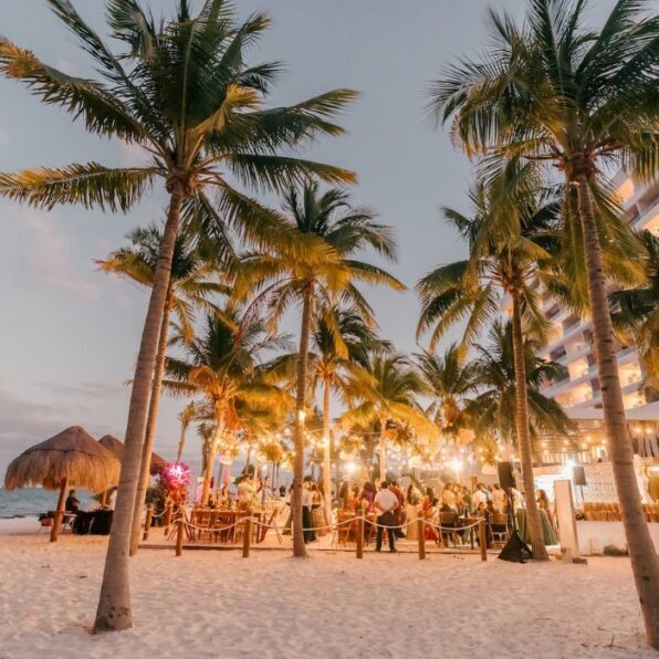 Outdoor beach event at sunset with people gathered under palm trees and string lights, near a thatched hut and a multi-story building.