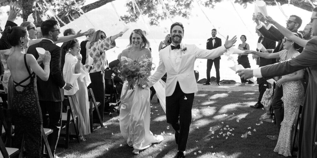 A newlywed couple walks down the aisle while guests cheer and toss petals. The event takes place outdoors under a tree canopy.