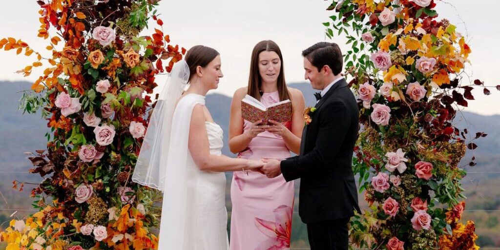 A bride and groom hold hands during an outdoor wedding ceremony, standing under a floral arch with an officiant reading from a book.