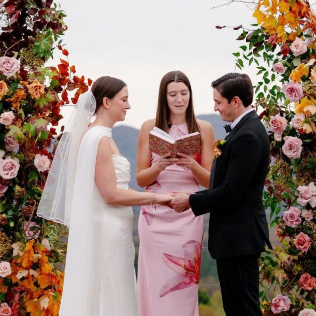 A bride and groom hold hands during an outdoor wedding ceremony, standing under a floral arch with an officiant reading from a book.