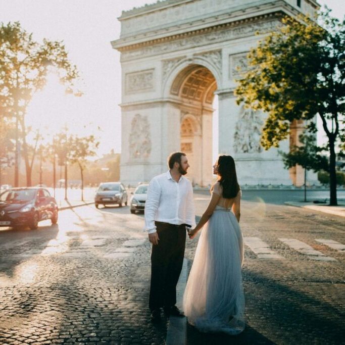 A couple holding hands while standing in front of the Arc de Triomphe in Paris.