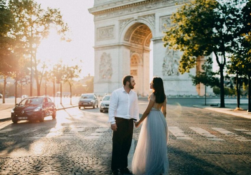 A couple holding hands while standing in front of the Arc de Triomphe in Paris.