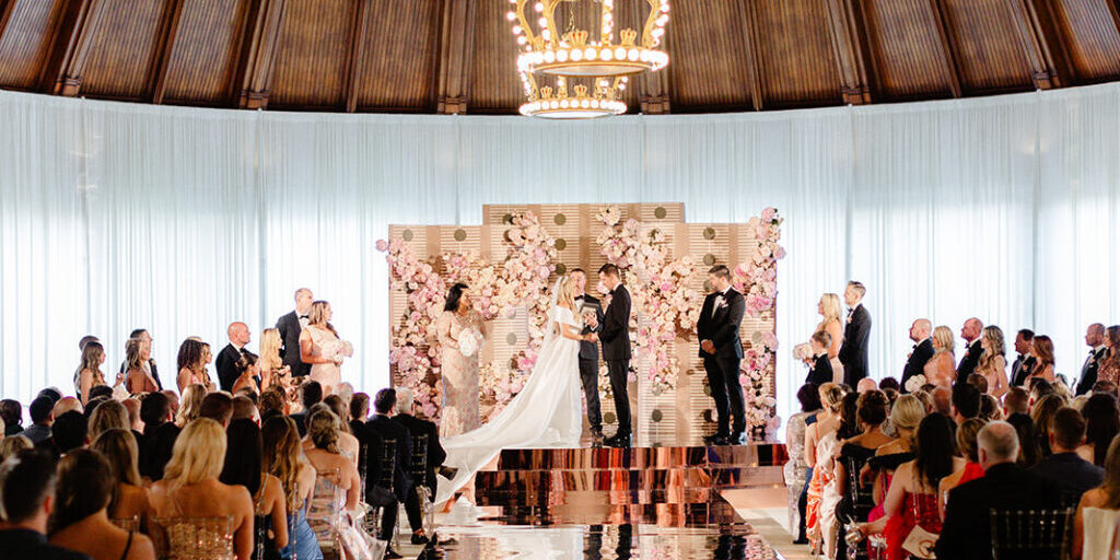 A wedding ceremony inside a domed room with a large chandelier overhead. The couple stands at the altar surrounded by a backdrop of pink flowers, with guests seated on either side.