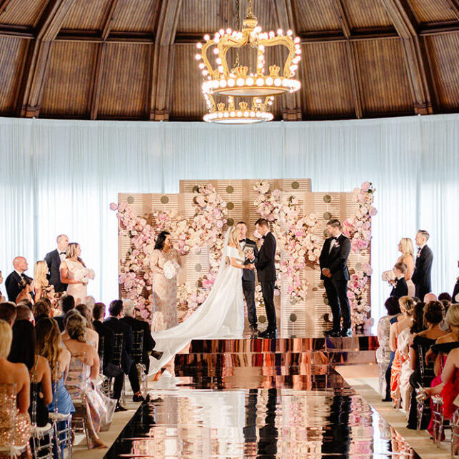 A wedding ceremony inside a domed room with a large chandelier overhead. The couple stands at the altar surrounded by a backdrop of pink flowers, with guests seated on either side.