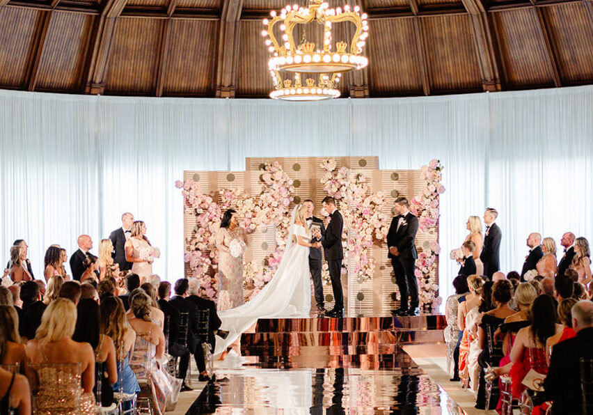 A wedding ceremony inside a domed room with a large chandelier overhead. The couple stands at the altar surrounded by a backdrop of pink flowers, with guests seated on either side.