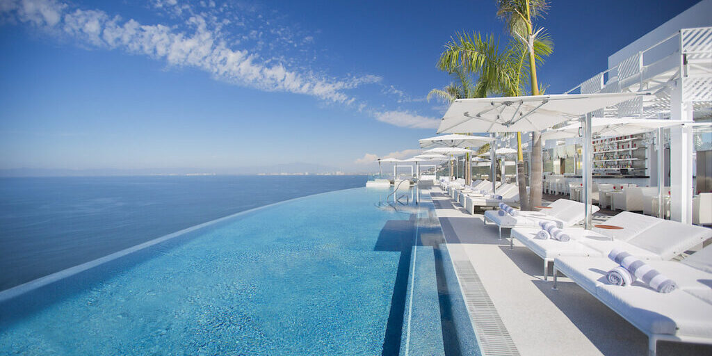 Infinity pool with clear blue water overlooks the ocean, lined with white lounge chairs, umbrellas, and palm trees under a clear sky.