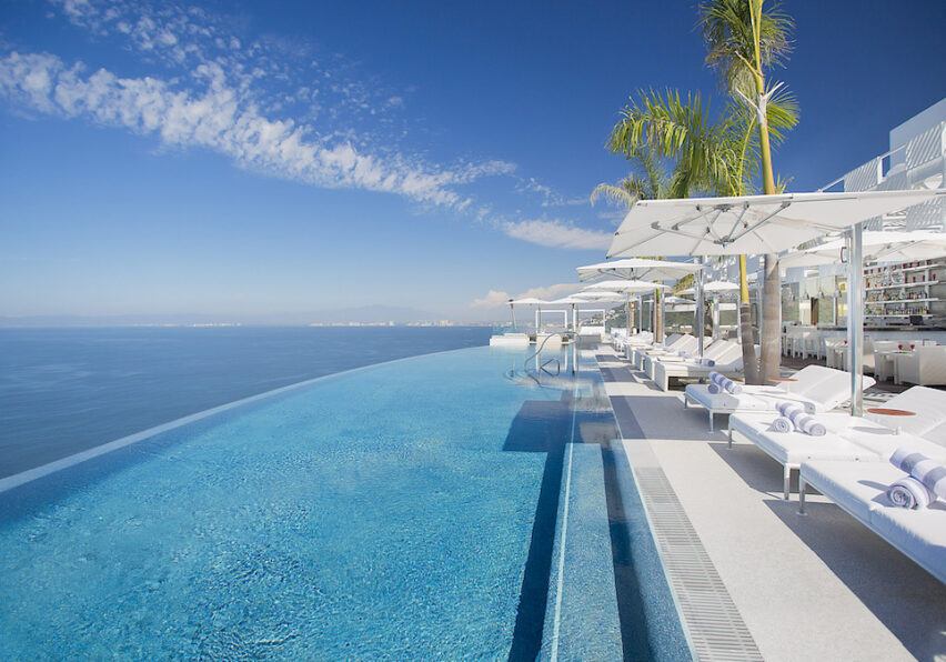 Infinity pool with clear blue water overlooks the ocean, lined with white lounge chairs, umbrellas, and palm trees under a clear sky.