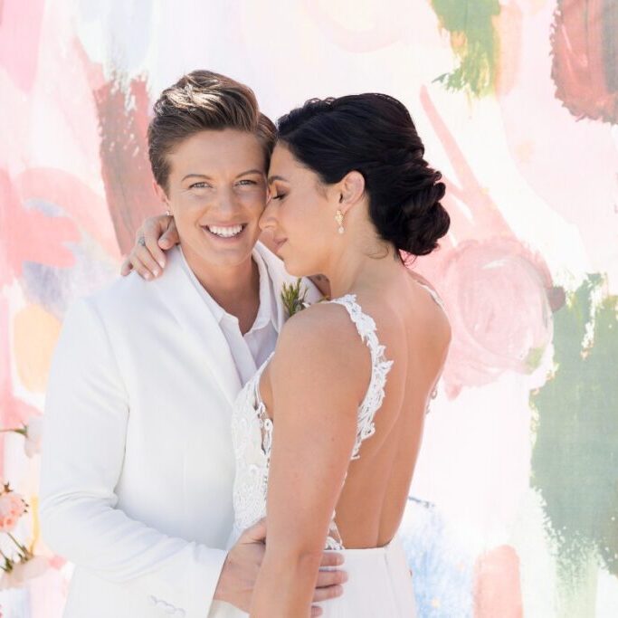 Two people in white wedding attire pose closely in front of a colorful backdrop, one smiling and the other resting their head on their shoulder. Floral arrangements are visible on the left side.