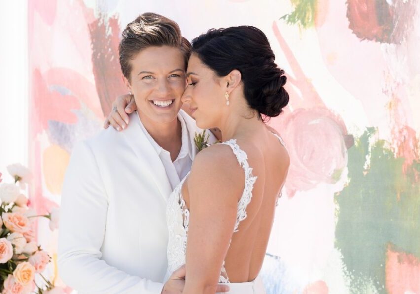 Two people in white wedding attire pose closely in front of a colorful backdrop, one smiling and the other resting their head on their shoulder. Floral arrangements are visible on the left side.