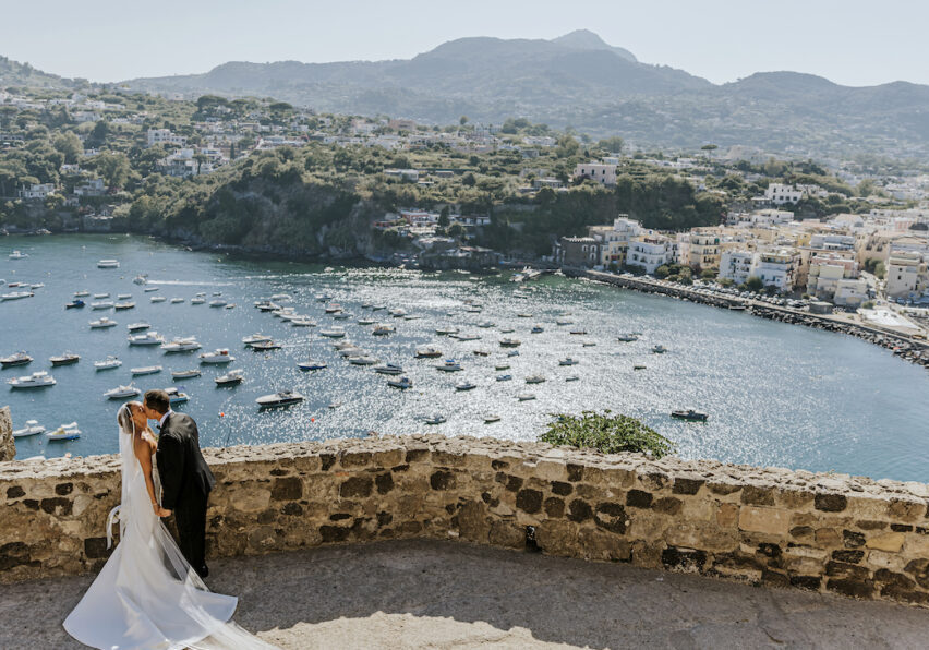 A bride and groom embrace on a stone terrace overlooking a coastal village and bay filled with boats on a sunny day.