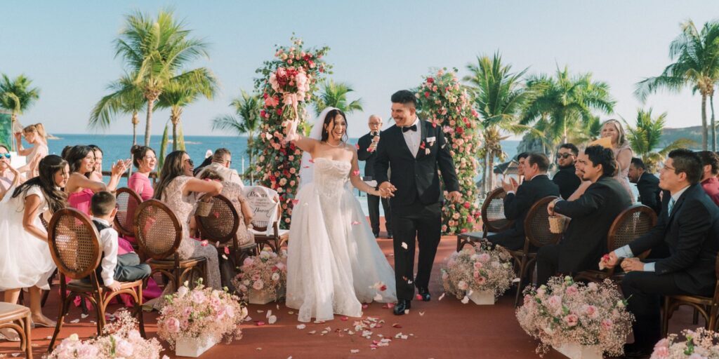 A bride and groom walk down an outdoor aisle lined with flowers, smiling and holding hands, surrounded by guests at a tropical wedding ceremony.