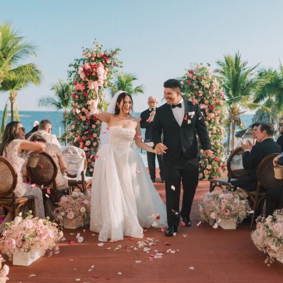 A bride and groom walk down an outdoor aisle lined with flowers, smiling and holding hands, surrounded by guests at a tropical wedding ceremony.