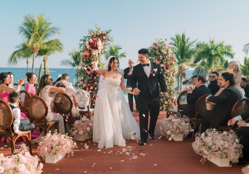 A bride and groom walk down an outdoor aisle lined with flowers, smiling and holding hands, surrounded by guests at a tropical wedding ceremony.