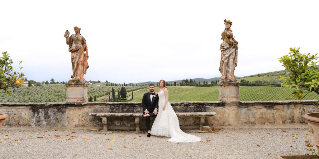 A bride and groom sit on a stone bench between two statues, with a scenic green landscape and cloudy sky in the background.