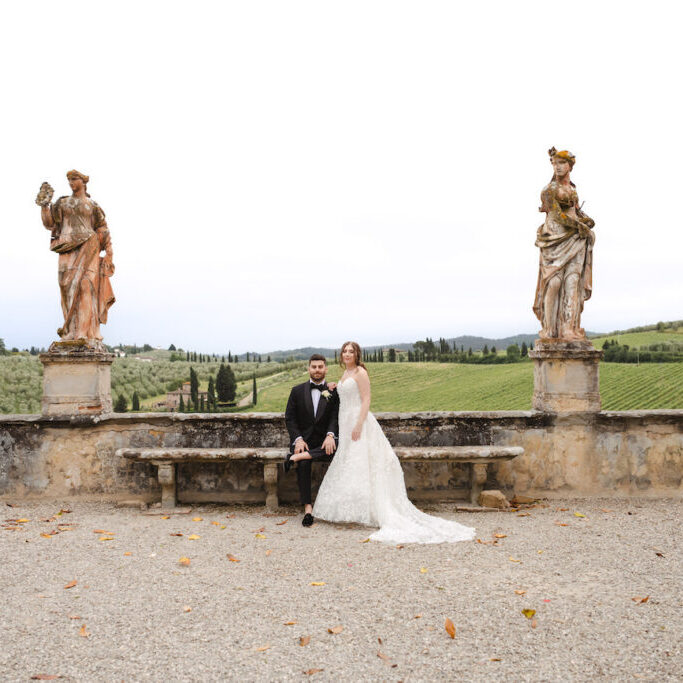 A bride and groom sit on a stone bench between two statues, with a scenic green landscape and cloudy sky in the background.