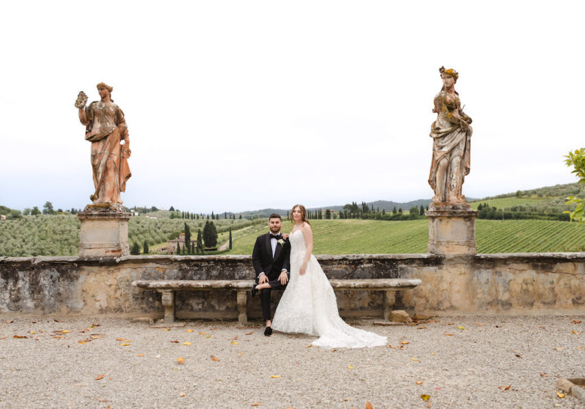 A bride and groom sit on a stone bench between two statues, with a scenic green landscape and cloudy sky in the background.
