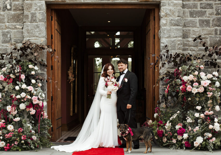 Bride and groom stand together with two dogs on a red carpet in front of a stone building, surrounded by large floral arrangements.
