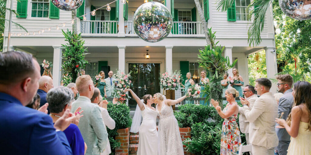 Two brides kiss and raise their arms in celebration at an outdoor wedding ceremony, surrounded by applauding guests, with a large disco ball and greenery in the background.