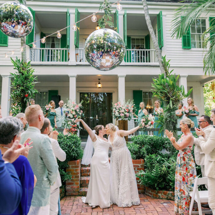 Two brides kiss and raise their arms in celebration at an outdoor wedding ceremony, surrounded by applauding guests, with a large disco ball and greenery in the background.