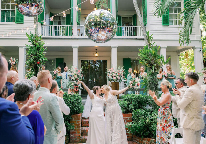 Two brides kiss and raise their arms in celebration at an outdoor wedding ceremony, surrounded by applauding guests, with a large disco ball and greenery in the background.