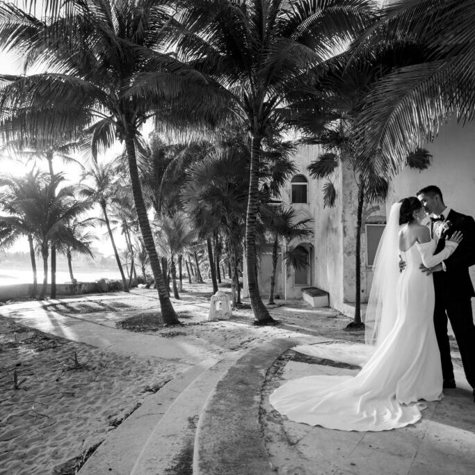 A bride and groom embrace under palm trees beside a sandy beach near a building, with sunlight filtering through the trees in this black and white photo.