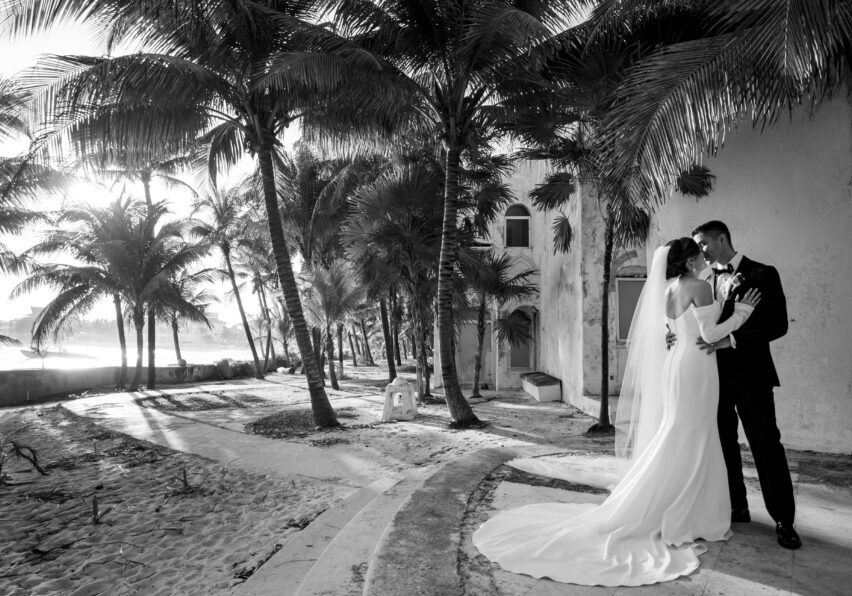 A bride and groom embrace under palm trees beside a sandy beach near a building, with sunlight filtering through the trees in this black and white photo.