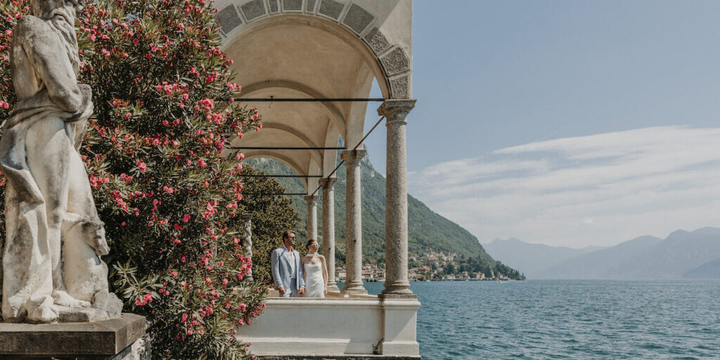 A couple stands under stone arches beside a flowering bush, overlooking a lake with mountains and a distant village in the background.