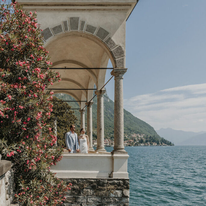 A couple stands under stone arches beside a flowering bush, overlooking a lake with mountains and a distant village in the background.