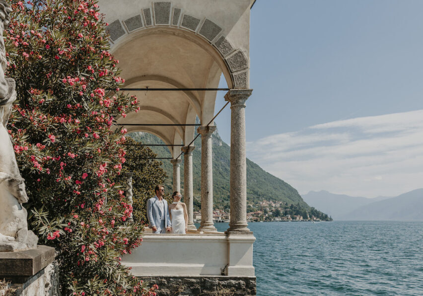 A couple stands under stone arches beside a flowering bush, overlooking a lake with mountains and a distant village in the background.