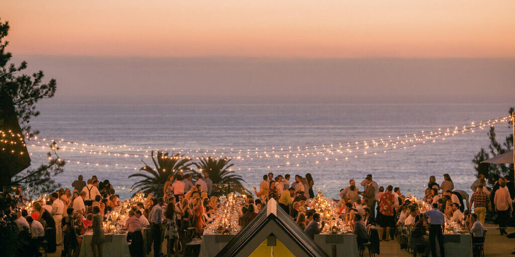 A large outdoor gathering at sunset near the ocean, with string lights overhead and tables full of people dining.