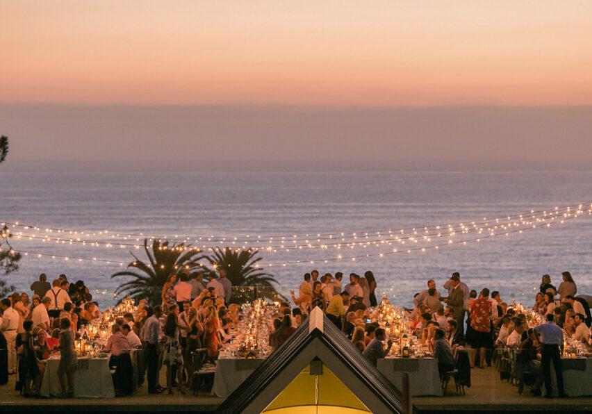 A large outdoor gathering at sunset near the ocean, with string lights overhead and tables full of people dining.