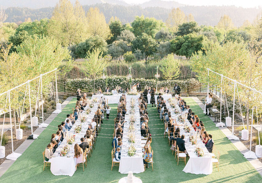 Outdoor wedding reception with long banquet tables, guests seated, string lights overhead, and lush greenery in the background.