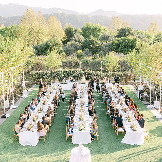 Outdoor wedding reception with long banquet tables, guests seated, string lights overhead, and lush greenery in the background.