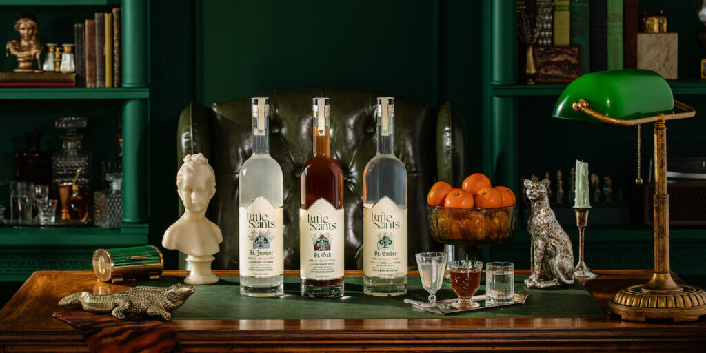 Four bottles of liquor on a wooden desk with glasses, a green desk lamp, books, a bust, a silver dog figurine, and a bowl of oranges in a richly decorated study.