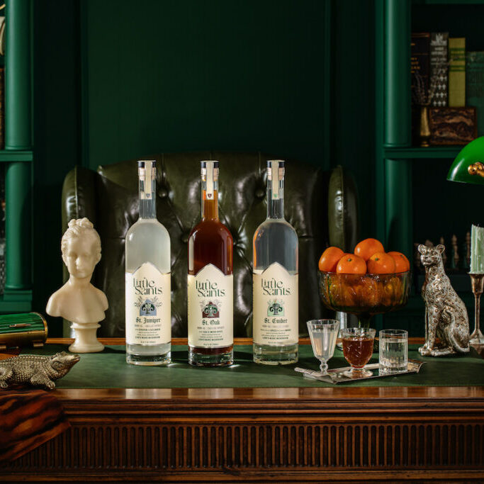 Four bottles of liquor on a wooden desk with glasses, a green desk lamp, books, a bust, a silver dog figurine, and a bowl of oranges in a richly decorated study.