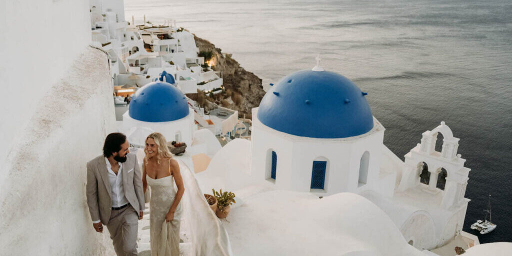 A couple in formal attire walks up white stairs beside blue-domed buildings, overlooking the sea in Santorini, Greece, at sunset.