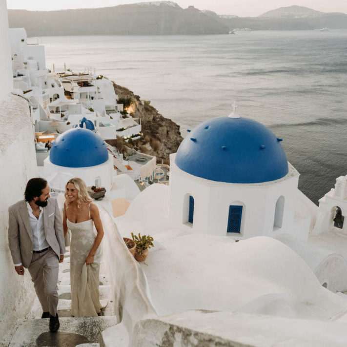 A couple in formal attire walks up white stairs beside blue-domed buildings, overlooking the sea in Santorini, Greece, at sunset.