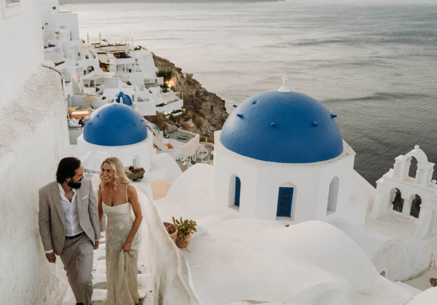 A couple in formal attire walks up white stairs beside blue-domed buildings, overlooking the sea in Santorini, Greece, at sunset.