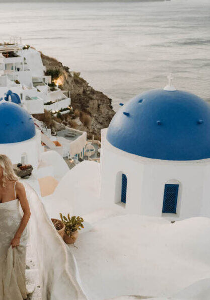 A couple in formal attire walks up white stairs beside blue-domed buildings, overlooking the sea in Santorini, Greece, at sunset.