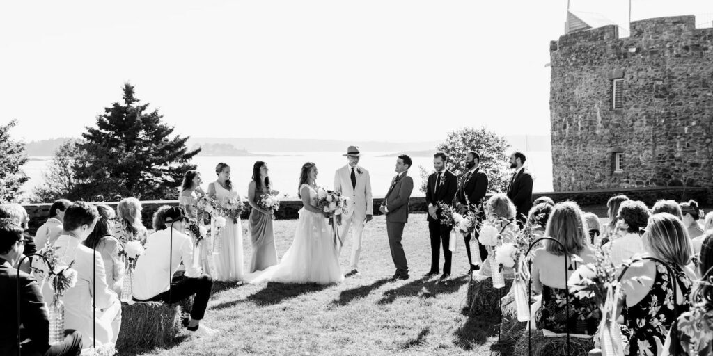 A wedding ceremony takes place outdoors, with the bride and groom facing each other, attendants standing by, and guests seated on either side.
