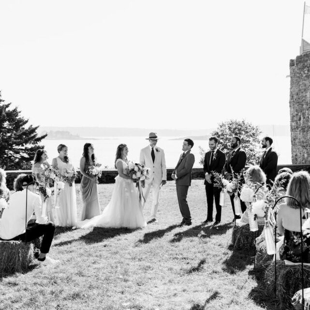 A wedding ceremony takes place outdoors, with the bride and groom facing each other, attendants standing by, and guests seated on either side.
