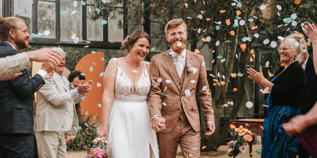A bride and groom walk hand in hand down the aisle while guests throw confetti and applaud at an indoor wedding ceremony.