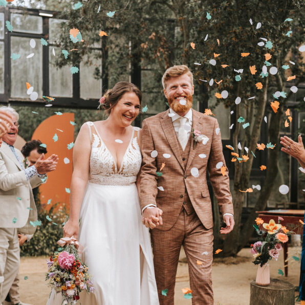A bride and groom walk hand in hand down the aisle while guests throw confetti and applaud at an indoor wedding ceremony.