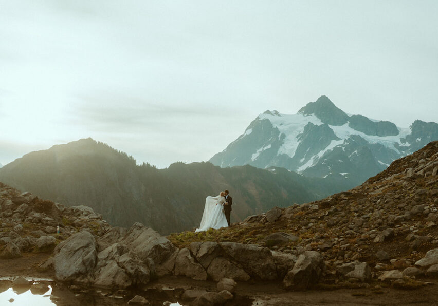 A couple in wedding attire stands on a rocky landscape with a snow-capped mountain and cloudy sky in the background.