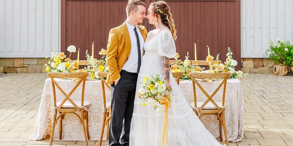 A couple stands in front of a decorated wedding table. The man wears a mustard jacket; the woman is in a white lace dress holding a bouquet. The backdrop features a wooden barn door.